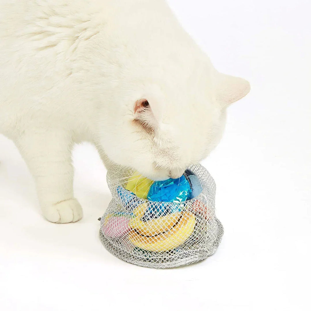 White cat sniffing a mesh bag filled with colorful cat toys including a banana and blue ball by zigly