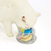 White cat sniffing a mesh bag filled with colorful cat toys including a banana and blue ball by zigly