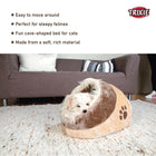 Small white dog resting inside a soft brown cave-shaped pet bed on a carpet in living room with couch and cushions zigly